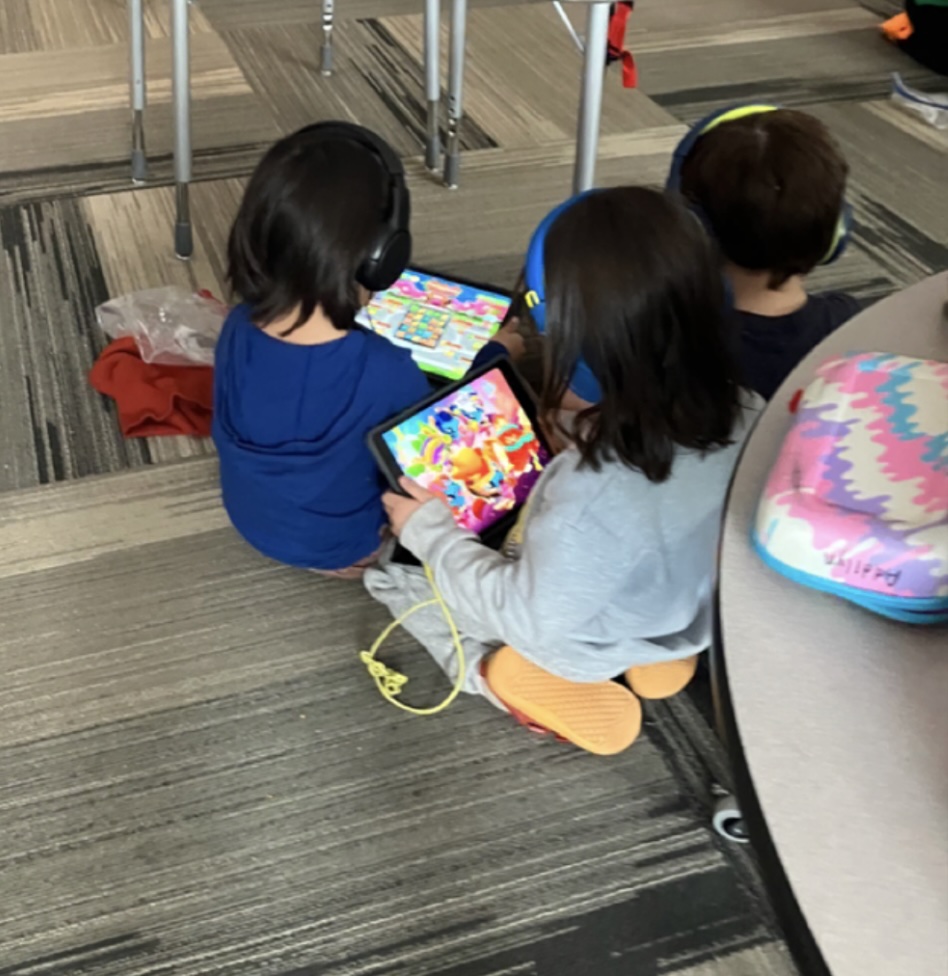 Three young children sitting on a classroom carpet wearing headphones, eyes locked on iPads displaying brightly colored game-like content.