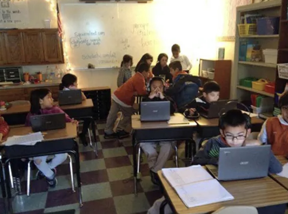 Elementary students seated at individual desks, each with an open Chromebook laptop, in a dimly lit classroom.