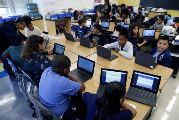 Students in a brightly lit classroom seated at group tables, each with a laptop open in front of them.